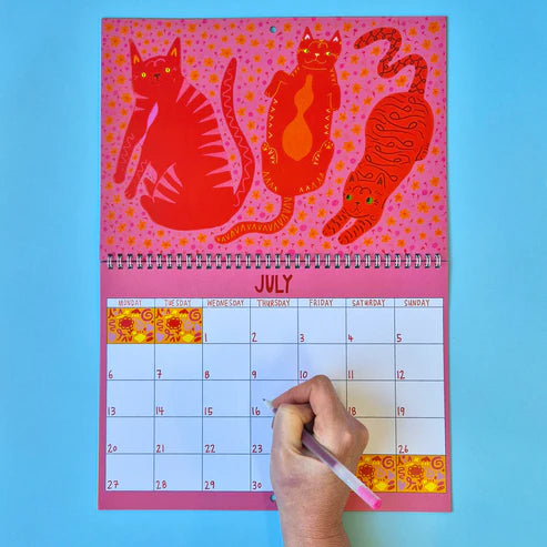 Calendar with red cat illustrations on a pink background, held by a hand with a pen.