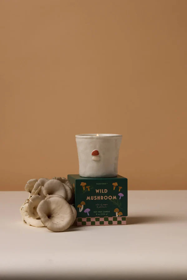 Candle in a white holder with a green box labeled 'Wild Mushroom' on a beige surface and background.