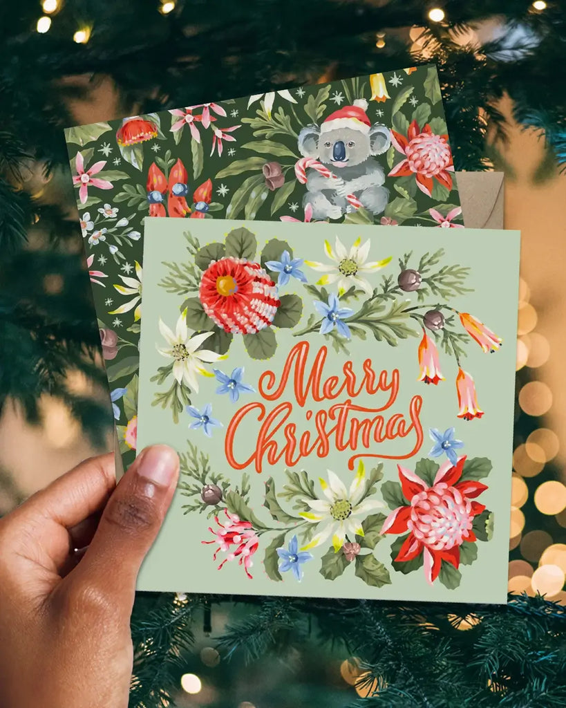 Hand holding a Christmas card with floral design and 'Merry Christmas' text, surrounded by festive lights and decorations.