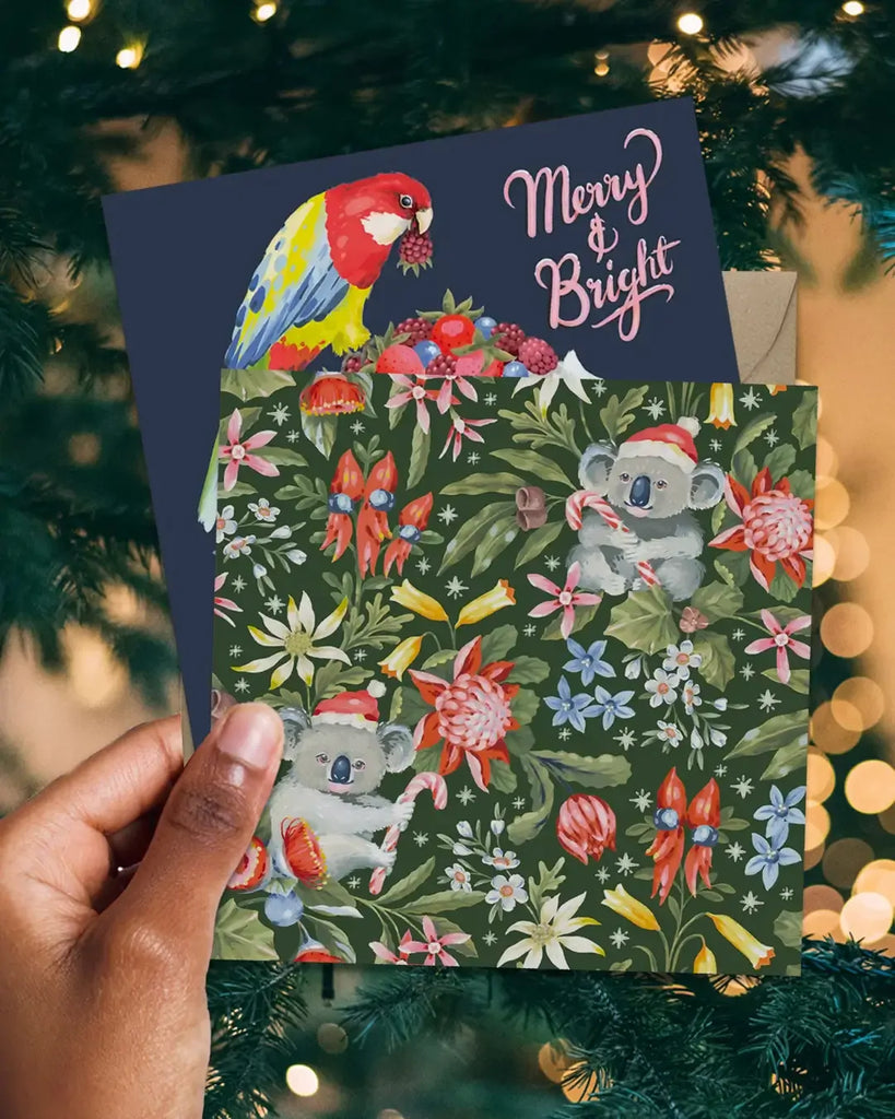 Christmas card with koalas, parrots, and flowers held in front of a decorated Christmas tree.