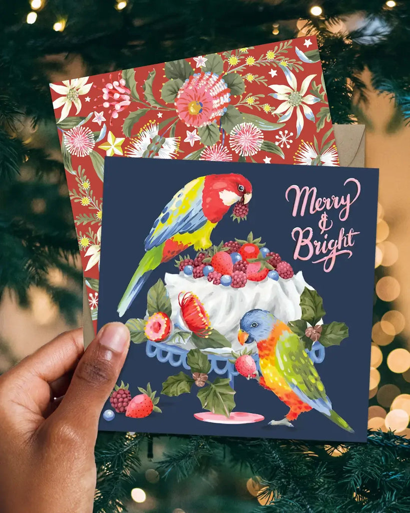 Hand holding a Christmas card with parrots and festive design against a blurred Christmas tree background.