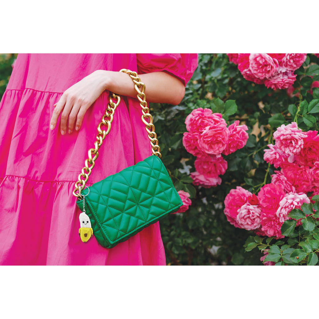 Green quilted handbag with gold chain strap and banana key chain held by a person in a pink dress, with pink flowers in the background.