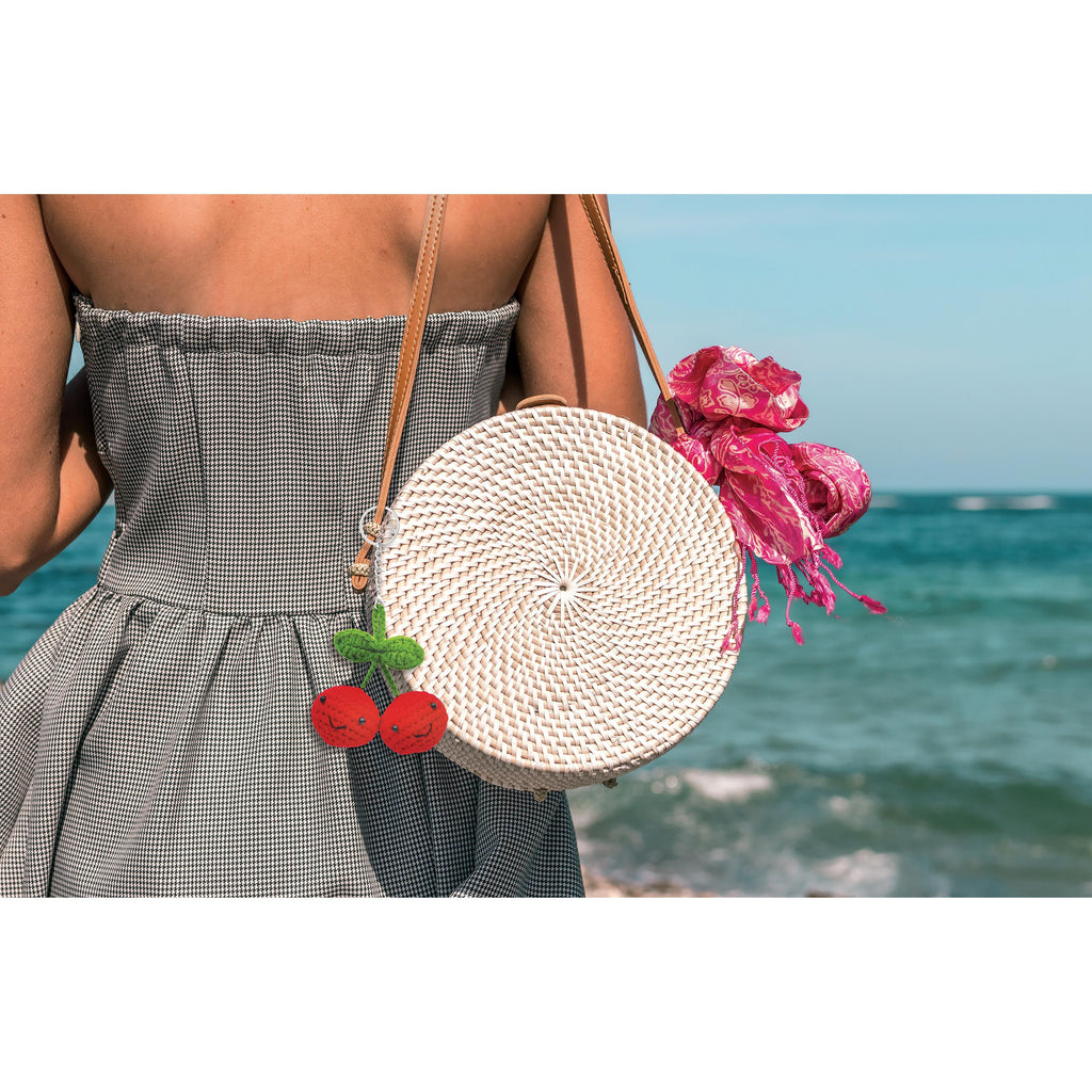 Person holding a woven handbag with floral and cherry keychain by the ocean.