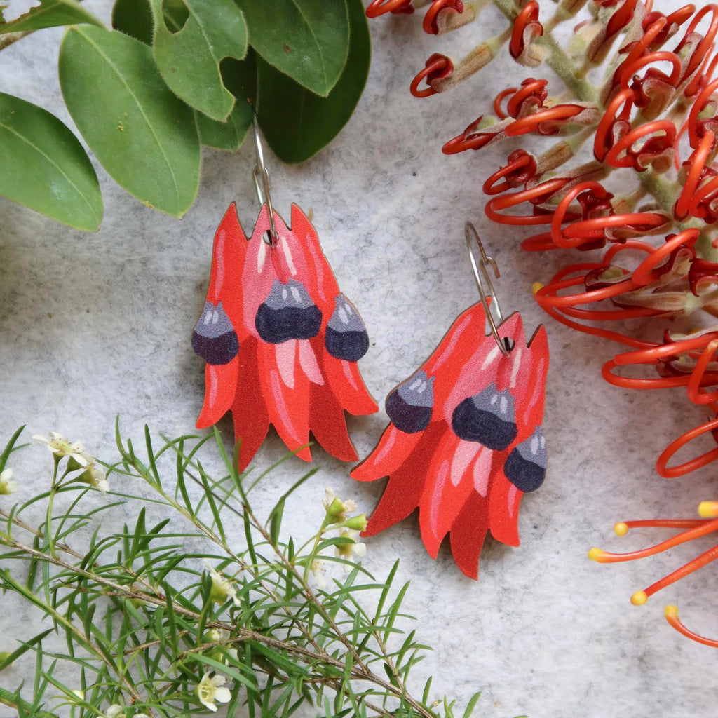Red floral earrings with black centers on a textured surface with green and orange plants.