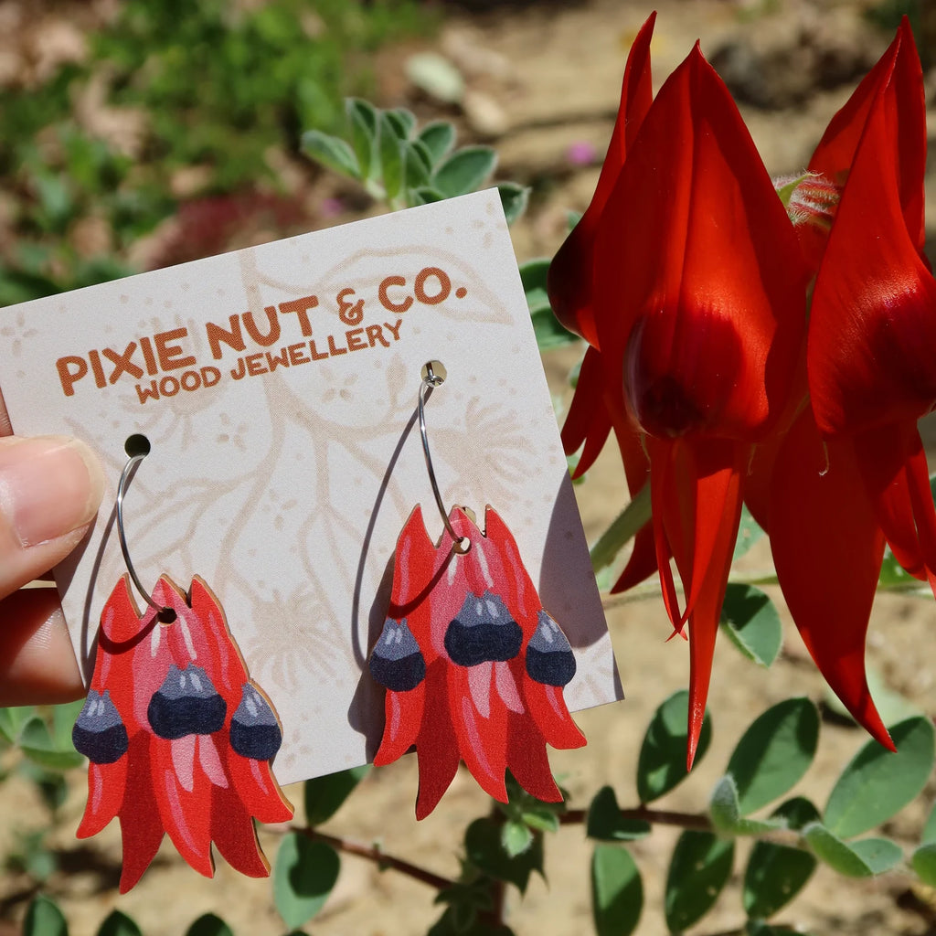 Red flower-shaped earrings on a Pixie Nut & Co. card with a red flower in the background